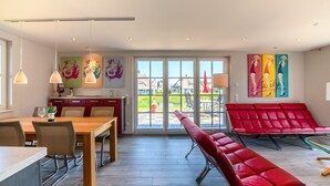 A modern dining area with a wooden table, chairs, and a red bench. There are colorful artworks on the walls and a view of the outdoors through large windows. - Reetdachhaus am Bodden mit Sauna (Fuhlendorf)