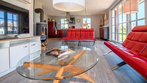 A modern living room with a glass coffee table, red leather sofa, and a large window providing a view of the outdoors. - Reetdachhaus am Bodden mit Sauna (Fuhlendorf)