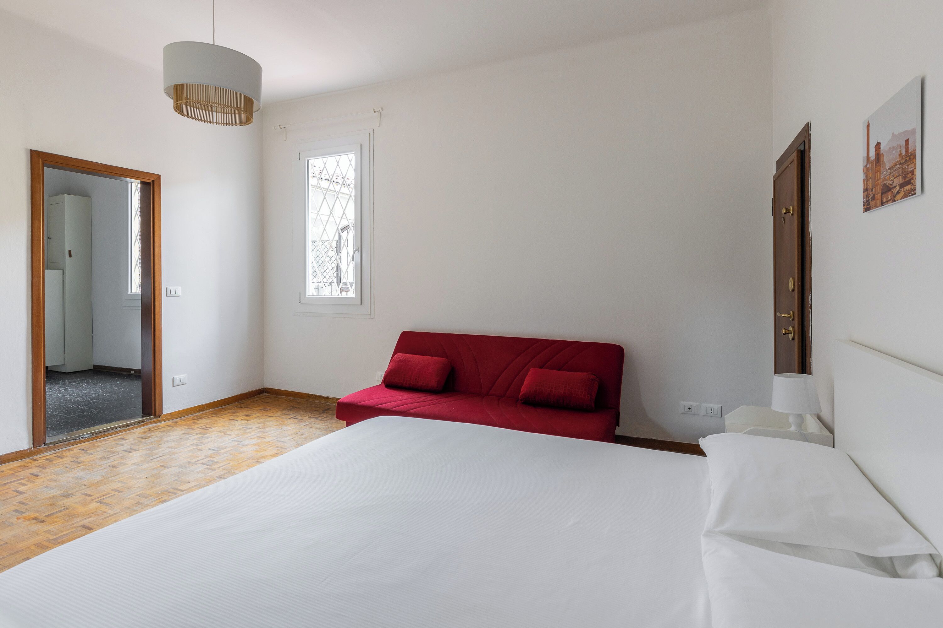 A bedroom with a large bed, a red sofa, a wooden floor, a window, and a door.