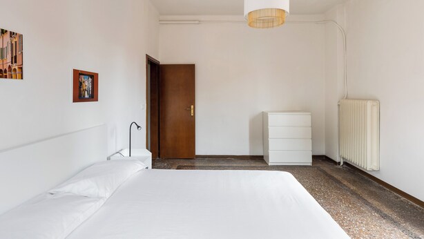 A bedroom with a bed, a white dresser, a door, a radiator, and a hanging light fixture. - Alessandrini Apartment alla Finestrella by Wonderful Italy (Bologna)