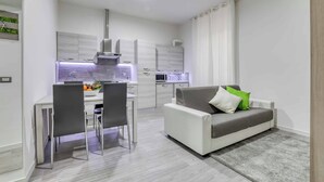 A modern kitchen with white cabinets, a dining area with a table and chairs, and a grey sofa with a green pillow. - Tourist Apartment Civitavecchia (Civitavecchia)
