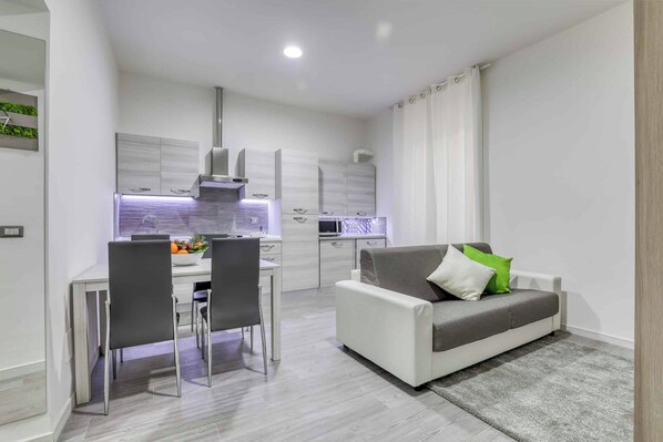 A modern kitchen with white cabinets, a dining area with a table and chairs, and a grey sofa with a green pillow. - Tourist Apartment Civitavecchia (Civitavecchia)