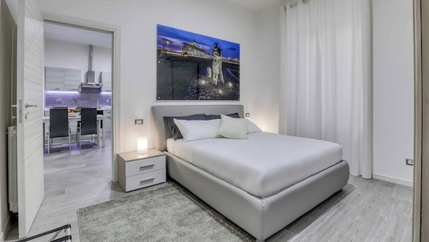 A modern bedroom with a large bed, a nightstand, a painting of a cityscape, and a view of a dining area. - Tourist Apartment Civitavecchia (Civitavecchia)