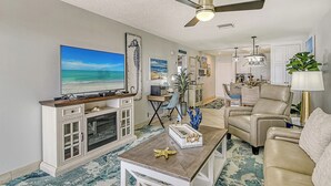 A living room with a large TV, a ceiling fan, a dining area, and a beige sofa.