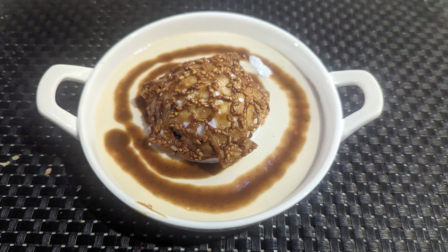 Una tazza bianca con manici che contiene un dessert guarnito con noci e un filo di sciroppo, posata su una superficie intrecciata.