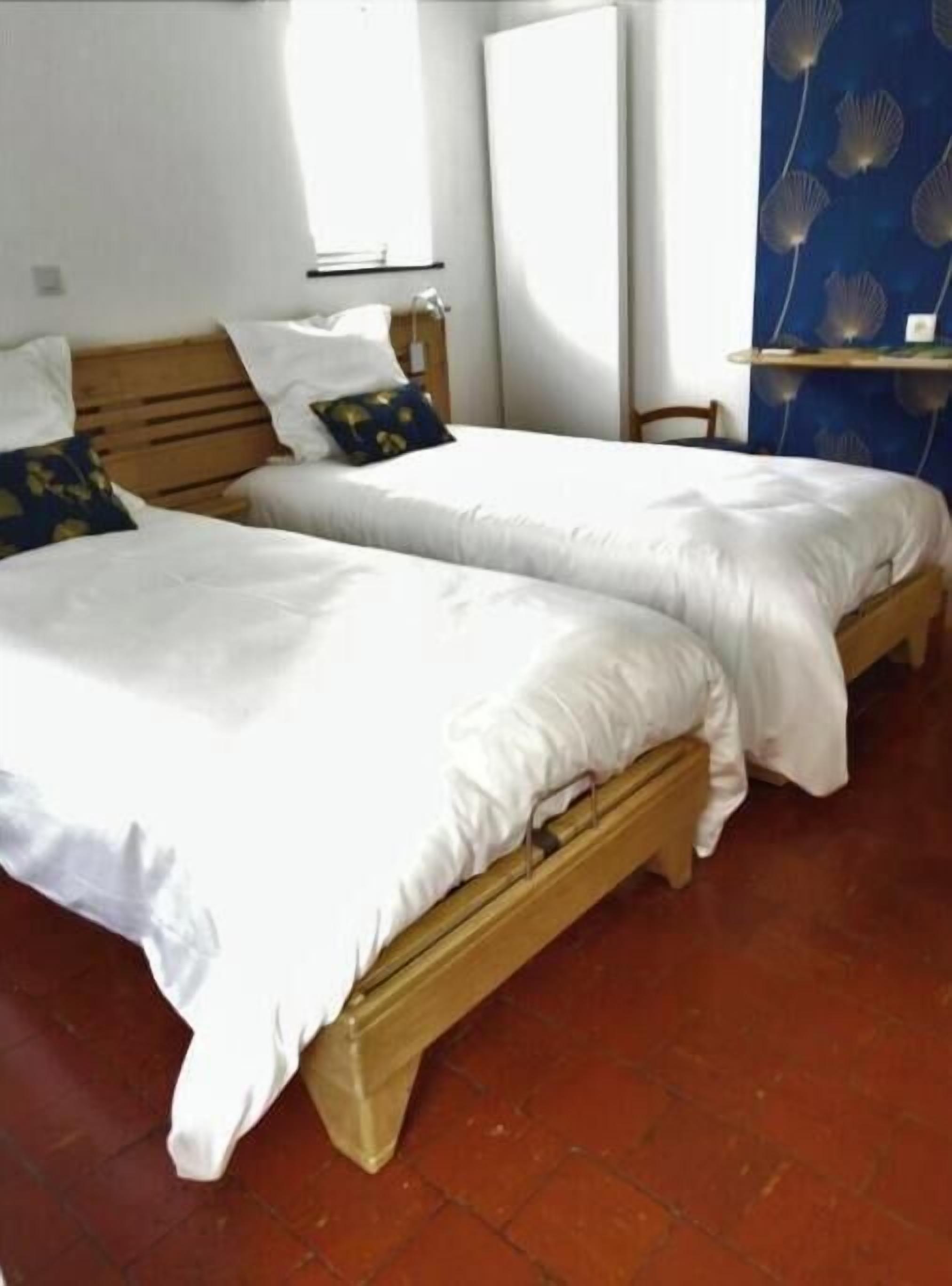 Une chambre avec deux lits en bois, recouverts de draps blancs, une table de chevet en bois, une étagère fixée au mur et une fenêtre avec des stores.