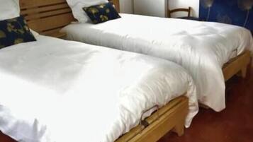 Une chambre avec deux lits en bois, recouverts de draps blancs, une table de chevet en bois, une étagère fixée au mur et une fenêtre avec des stores.