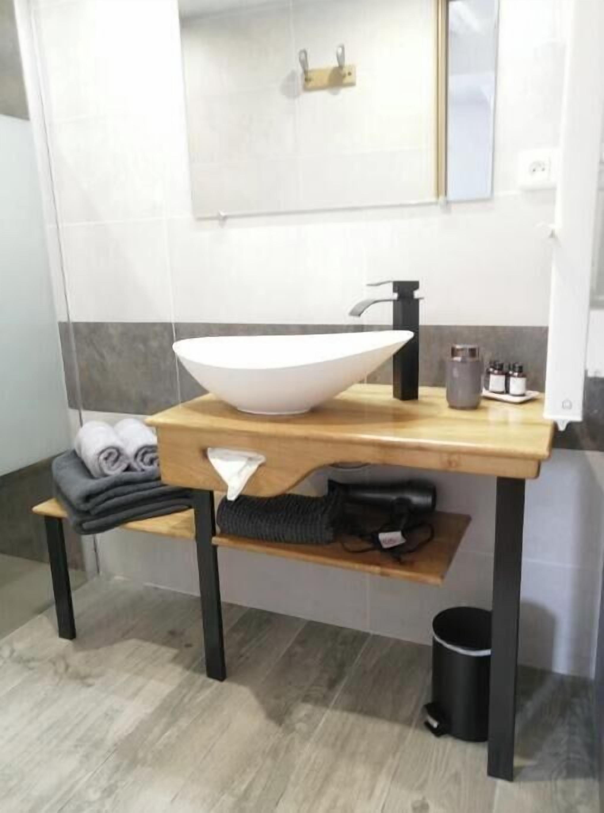 A modern bathroom with a large white vessel sink, a wooden countertop, a towel rack with towels, a small shelf, and a black trash can.