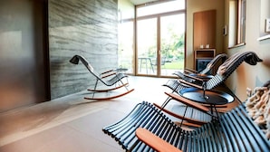 A modern interior with rocking chairs and a rattan sofa, large windows, and a kitchen area.