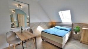 Un dormitorio con cama, escritorio y silla. Hay un espejo grande en la pared, una ventana y una planta en maceta.