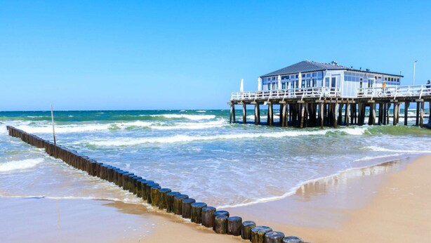 A beach with a wooden pier leading to a building with a balcony. - Comfortable apartment with a sea view (Ustronie Morskie)