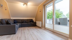 A modern living room with a sectional sofa, a large window with blinds, and a sloped ceiling. - Wohnung in St.Peter Ording (St.Peter Ording)