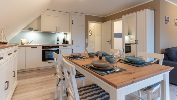 A modern kitchen with white cabinets, a wooden dining table set for four, and a visible oven. - Wohnung in St.Peter Ording (St.Peter Ording)