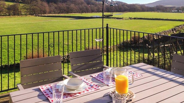 A dining table with chairs, a pitcher of orange juice, and a glass of water set on a patio with a view of a green field and stone wall.