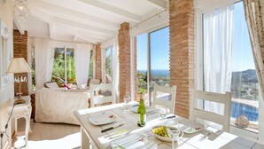 Une pièce lumineuse et ensoleillée, dotée d’une grande fenêtre donnant sur un paysage magnifique. On y trouve une table à manger dressée avec des assiettes, une bouteille de vin et un bol d’olives. La pièce est décorée de meubles blancs et comprend une colonne en briques.