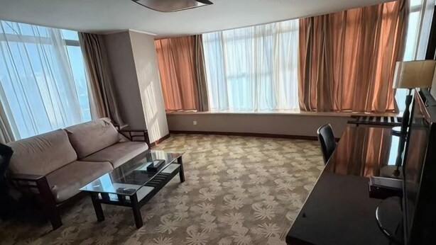 A hotel room with a sofa, a glass-top coffee table, a desk with a lamp, and large windows with curtains. - Chenguang Hotel (Xingtai)