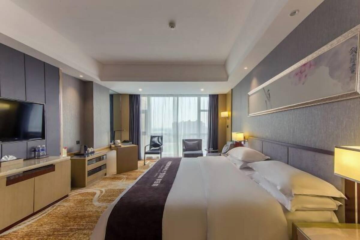 Habitación de hotel con una cama grande, televisión, escritorio y vista de la ciudad a través de amplias ventanas.