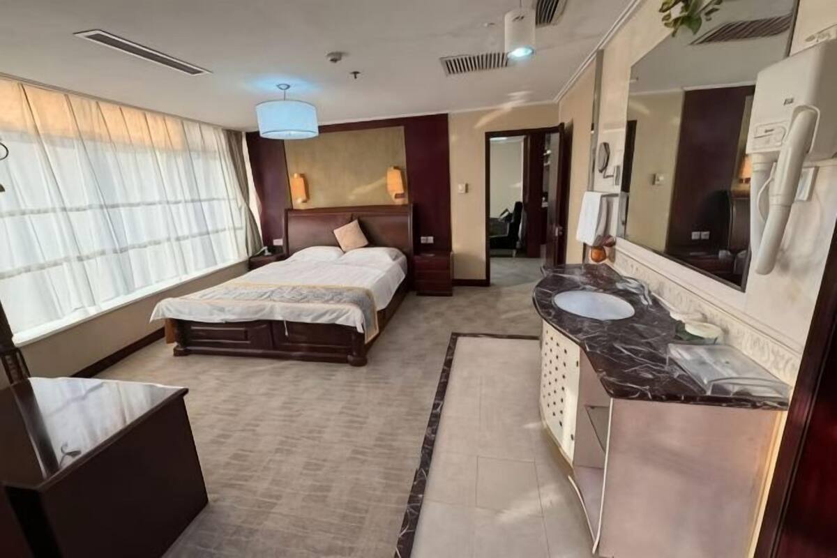 Habitación de hotel con cama, baño con lavamanos y espejo, y un ventanal grande con cortinas.