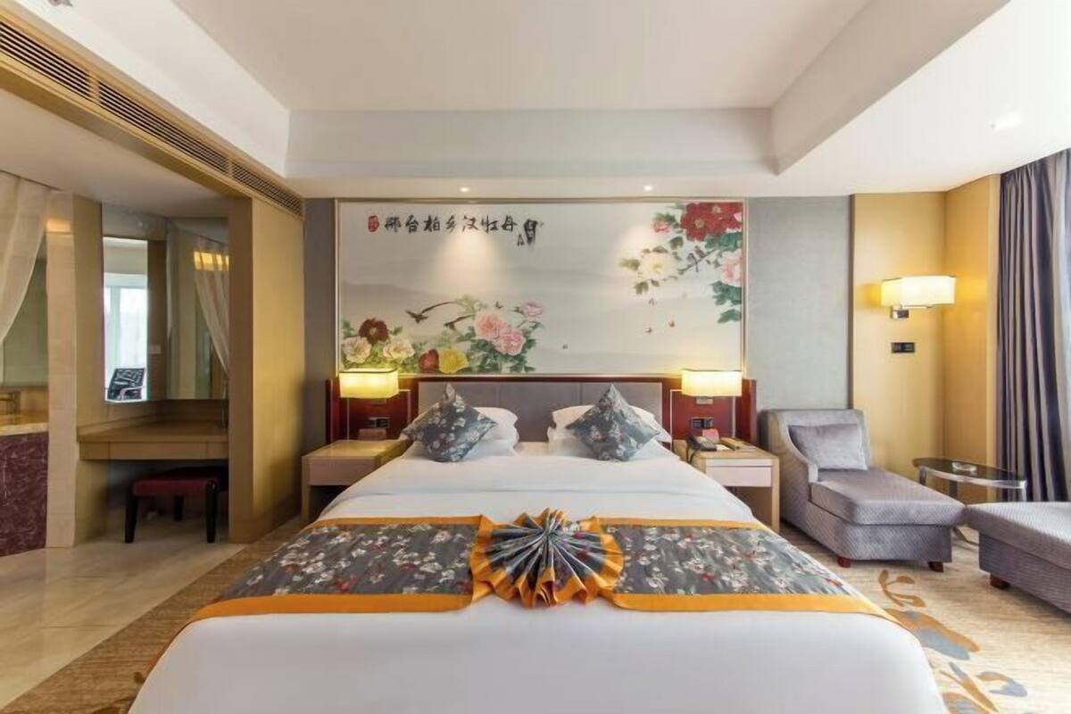 Habitación de hotel con una cama grande, dos mesitas de noche, una silla, un escritorio y un cuadro decorativo en la pared.