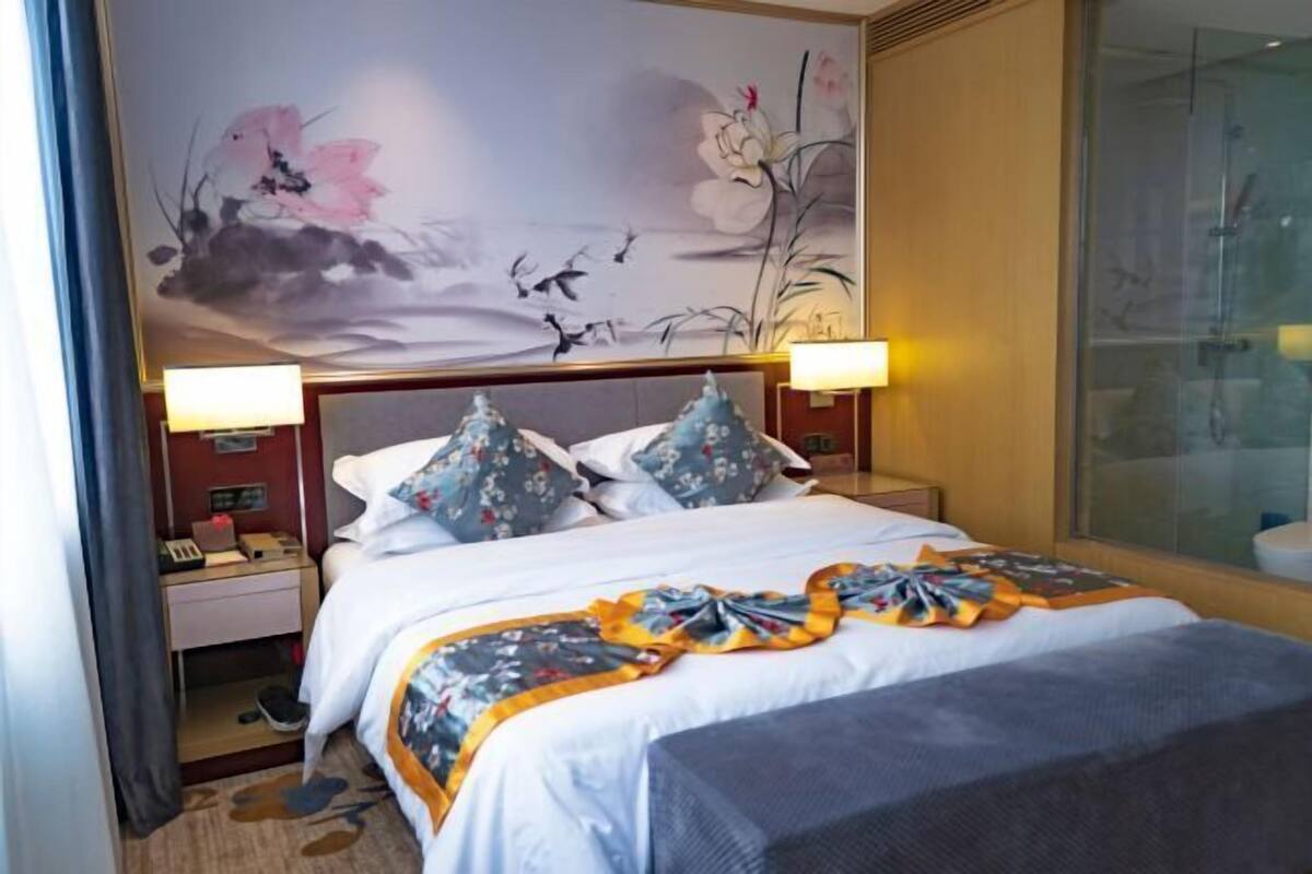 Una habitación de hotel con cama, almohadas, una mesita de noche, lámpara, un cuadro en la pared y un área de ducha con mampara de vidrio.