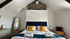 Un dormitorio con pared de piedra, una cama con ropa de cama blanca y dos almohadas amarillas, cabecero de madera, un espejo, una mesita de noche con lámpara y una silla con cojín amarillo.