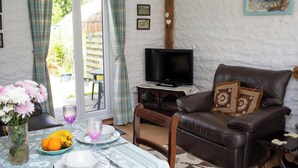 A living room with a TV, a brown leather sofa, a dining table with a vase of flowers, and a view through a glass door. - The Old Cottage, Aberporth (Glanrhyd)