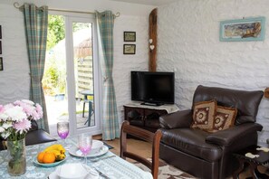 A living room with a television, a brown leather sofa, a dining table with a vase of flowers, and a view through a glass door. - The Old Cottage, Aberporth (Glanrhyd)