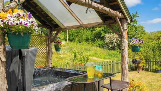 A hot tub with a wooden pergola, wicker chairs, and hanging flower pots. - The Old Cottage, Aberporth (Glanrhyd)