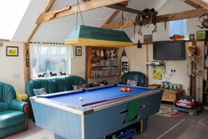A cozy living room with a pool table, green leather sofas, a bookshelf, and a television.