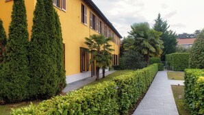 A yellow building with a paved pathway, tall coniferous trees, and a well-maintained hedge. - Medel (Appiano Gentile)