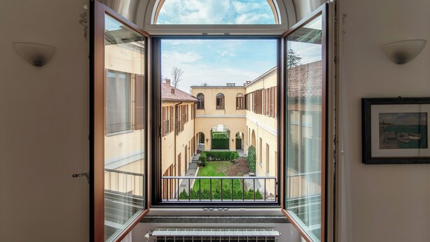 A view from a balcony with a glass door leading to a courtyard with buildings and greenery. - Medel (Appiano Gentile)