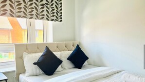 A bedroom with a tufted headboard, two pillows, a white bedspread, and a window with blinds. - Honeybee Haven Retreat - 3 Bed House (Ashford)