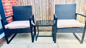 A patio area with two black wicker chairs and a small table, set against a wooden fence and brick wall. - Honeybee Haven Retreat - 3 Bed House (Ashford)