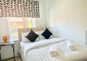 A hotel room with a bed, two pillows, a bedside table with a lamp, and a window with curtains. - Honeybee Haven Retreat - 3 Bed House (Ashford)