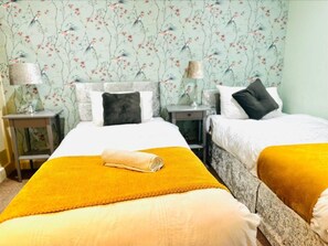 Ett hotellrum med två sängar, var och en med ett vitt täcke och en svart kudde. En gul handduk ligger på sängen i förgrunden. Rummet har blommig tapet och två sängbord med lampor.