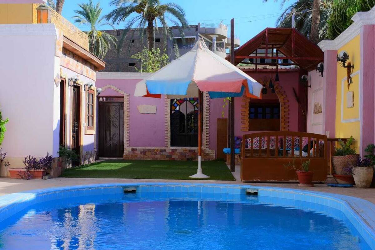 Uma piscina com área coberta, um pequeno jardim e um edifício com fachada rosa marcante.
