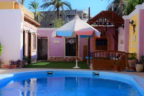 A swimming pool with a covered area, a small garden, and a building with a distinctive pink facade. - Sofia Guest House (Luxor)