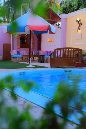 Un espace piscine doté d’une terrasse en bois, d’un parasol coloré et d’un coin salon avec des coussins à motifs.
