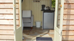 Une cabane en bois, dont la porte est ouverte, laissant entrevoir une petite cuisine, une chaise et un réfrigérateur compact.
