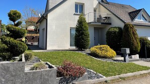Une maison moderne à deux étages, dotée d’un balcon, d’une cheminée et d’un jardin bien entretenu avec diverses plantes et un petit étang.