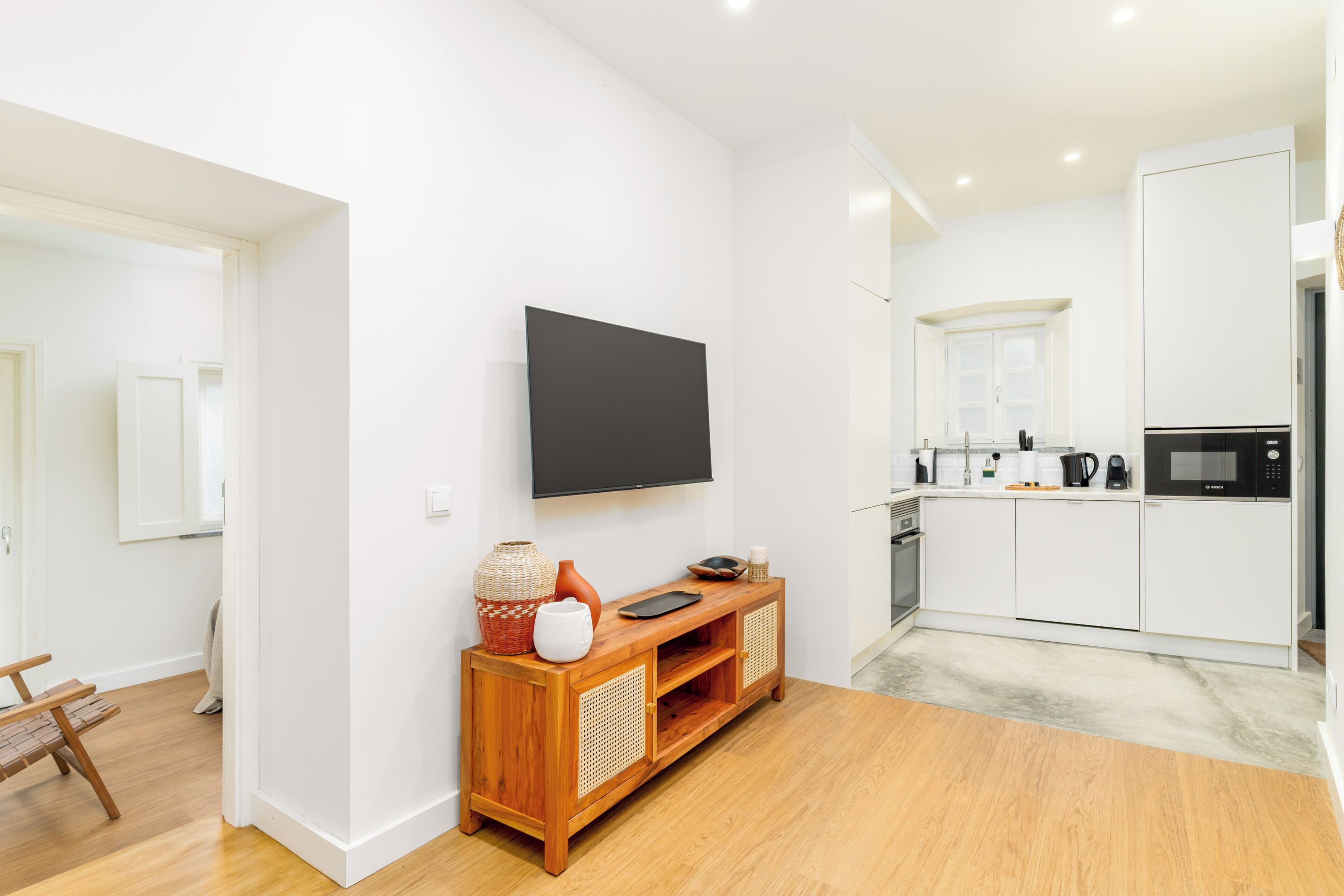 A modern, minimalist living room with a wooden entertainment unit, a flat-screen TV, and a kitchen area with white cabinetry and appliances.