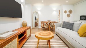 A modern living room with a wooden coffee table, a sofa with cushions, a TV mounted on the wall, a dining area with a table and chairs, and a kitchen area with a microwave.