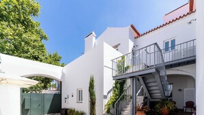 A white building with a staircase leading to a balcony, a green gate, and a paved courtyard.