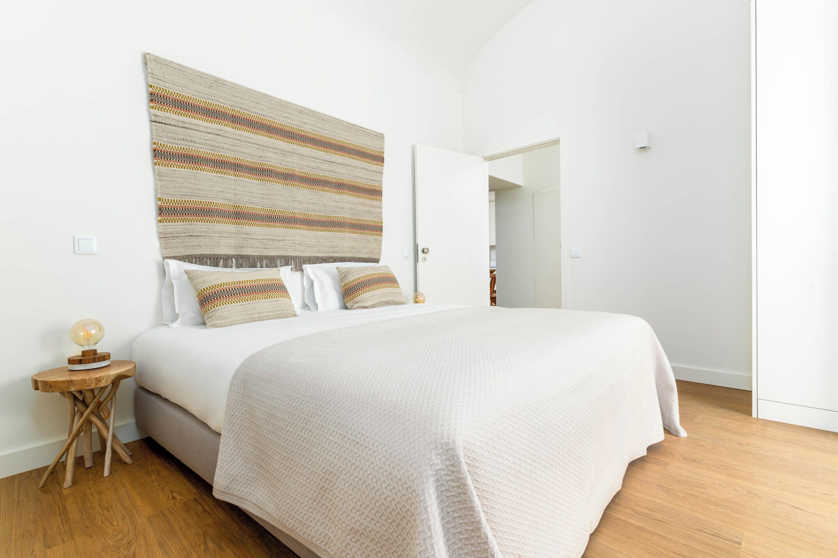 A bedroom with a large bed, a patterned wall hanging, a small wooden side table, and a door leading to another room.