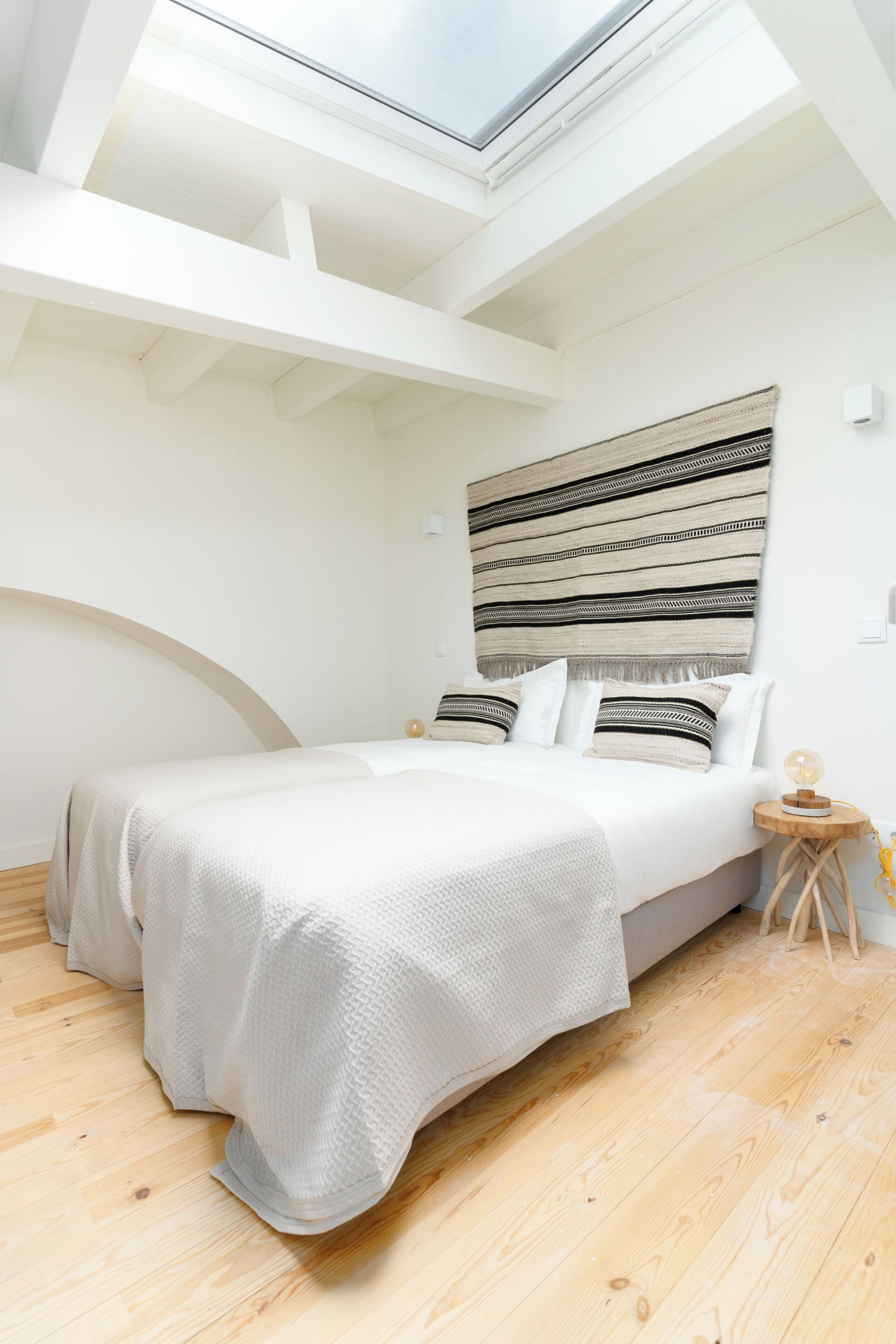 Une chambre avec un puits de lumière, un lit avec une literie blanche, un plancher en bois et une lampe fixée au mur.