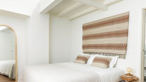 A bedroom with a large bed, a mirror, a wall hanging, and a small wooden side table with a lamp.