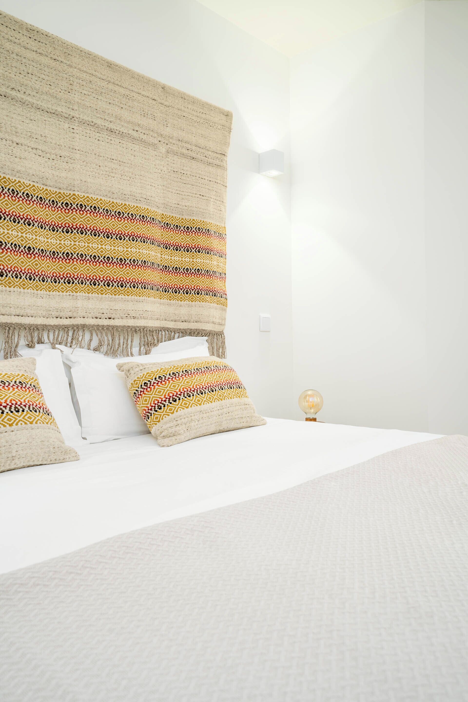 A neatly made bed with a white comforter and patterned pillows, a woven headboard, and a wall-mounted light fixture.
