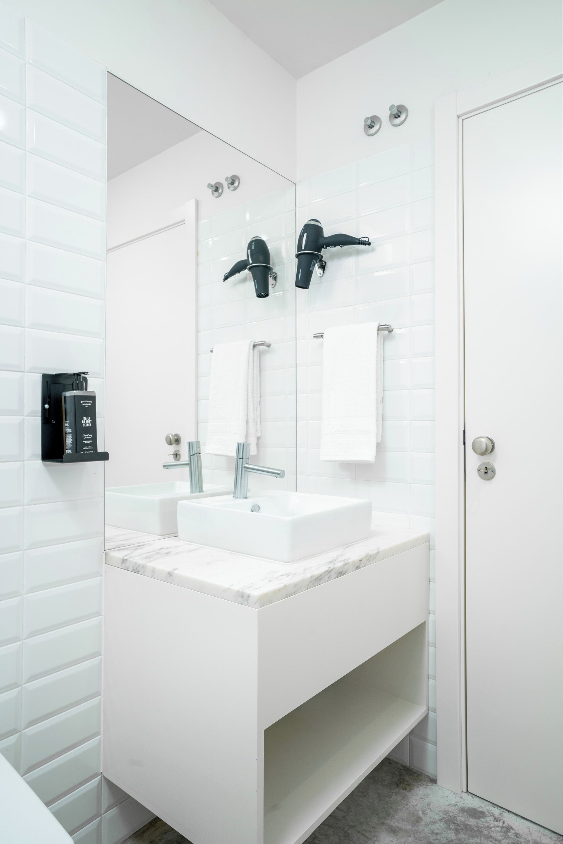 A modern bathroom with white tiled walls, a white vanity with a marble countertop, a large mirror, a towel rack, and a door with a keyhole.