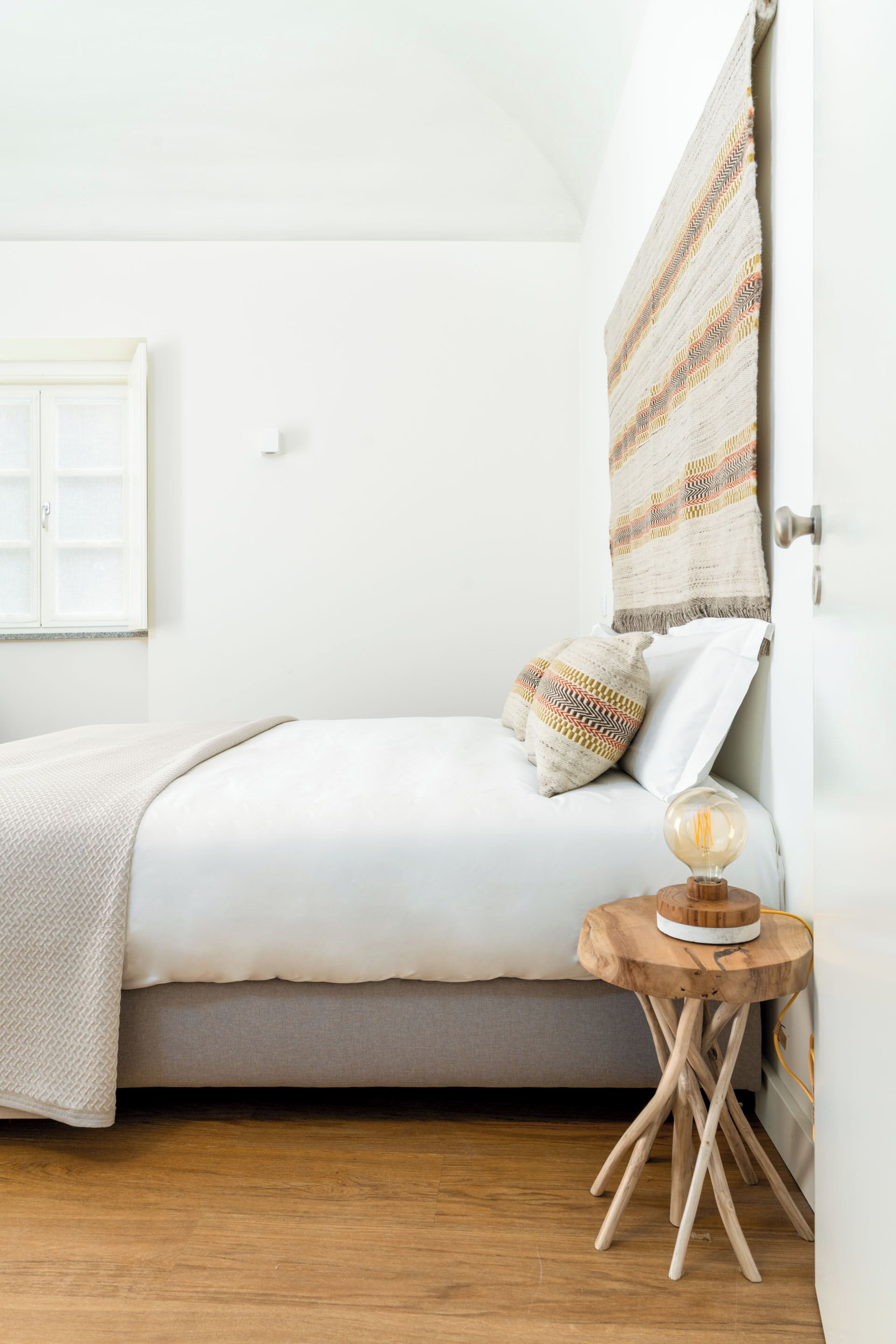 A bedroom with a bed, a bedside table with a lamp, a window with a view, and a wall with a patterned tapestry.