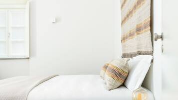 A bedroom with a bed, a bedside table with a lamp, a window with a view, and a wall with a patterned tapestry.